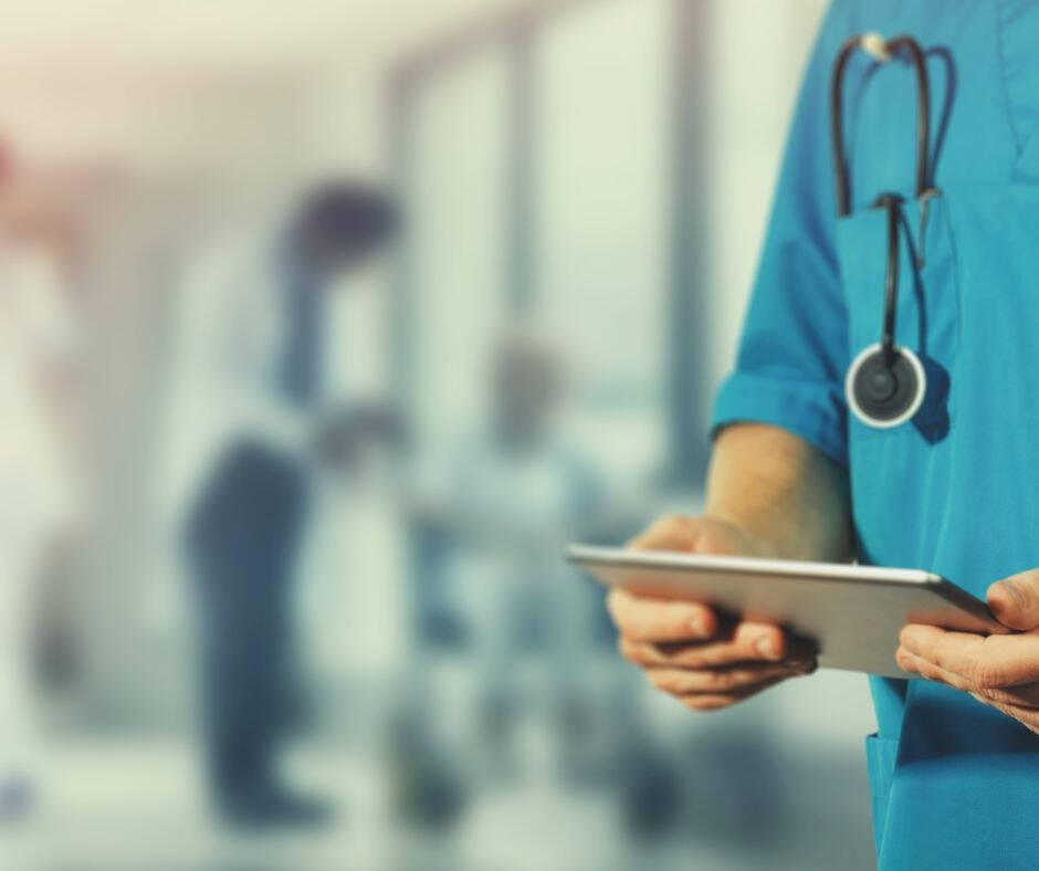 Medical professional wearing blue scrubs with a stethoscope around their neck and holding a tablet device. Blurred background suggests a hospital or clinic setting with staff and patient interaction beyond.